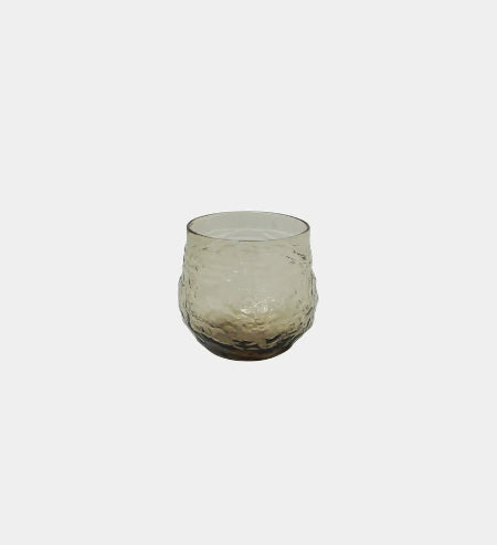 A small stemless glass tumbler with a textured, hammered finish, featuring a smoky translucent design that transitions from light gray at the top to a dark brown at the base, set against a solid light gray background.