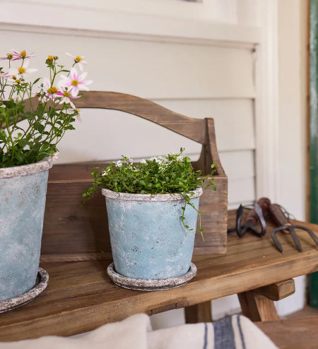 Two distressed light blue metal planters with white trim sit on a rustic wooden bench. One planter holds pink and white daisies, and the other holds green trailing foliage.