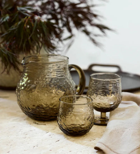 Set of brown glassware including a pitcher and two glasses on a textured surface with a plant in the background.